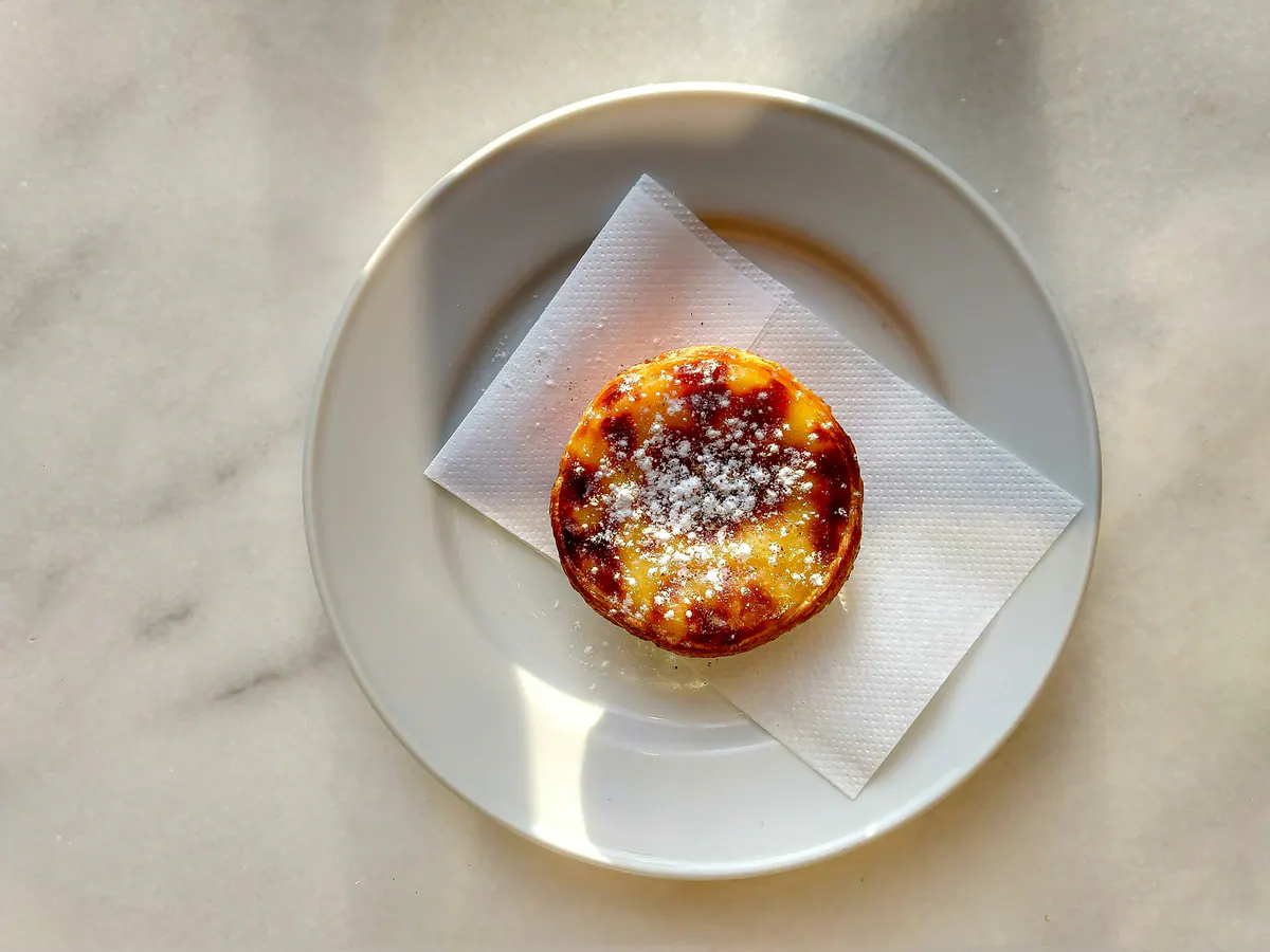 A pastel de nata dusted with cinnamon on a white plate, on a sunlit marble table