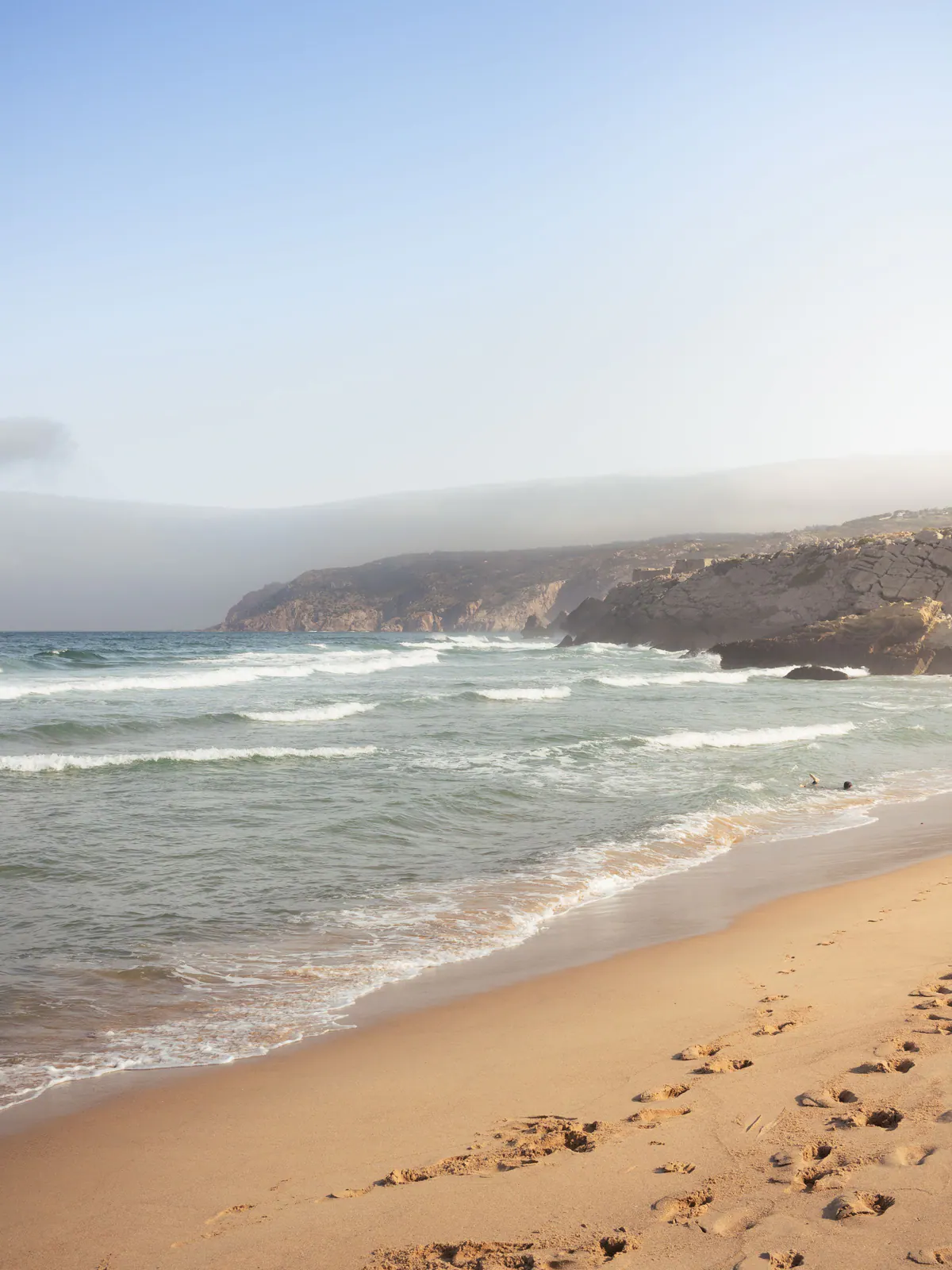 Praia Grande in golden light: footprints in the sand, gentle waves and misty cliffs