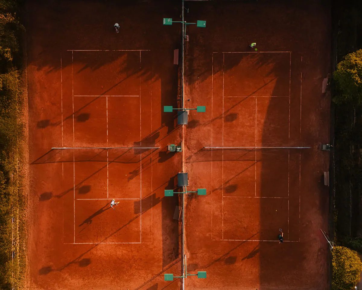 Clay tennis courts from above at golden hour, players casting long shadows