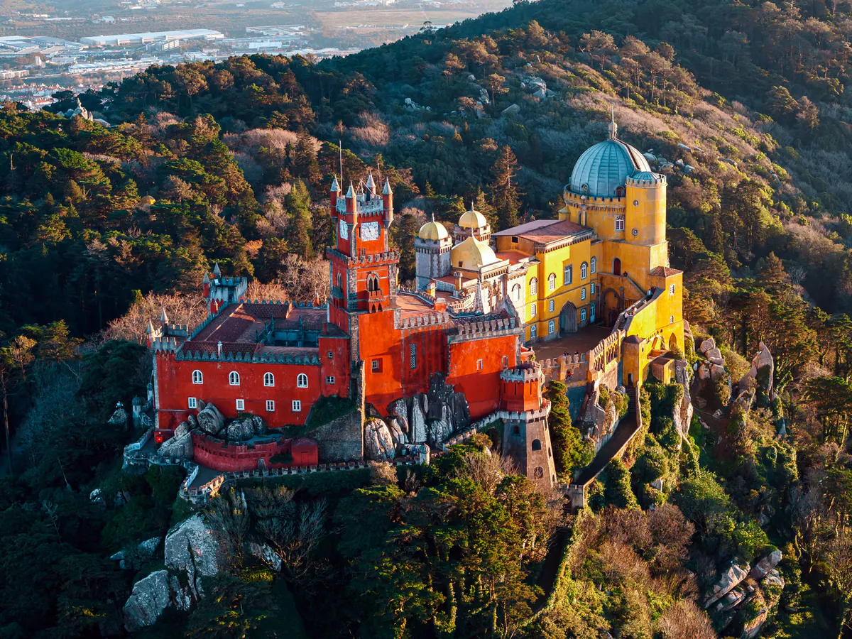 Aerial view of the red and yellow Pena Palace on its wooded peak in warm light